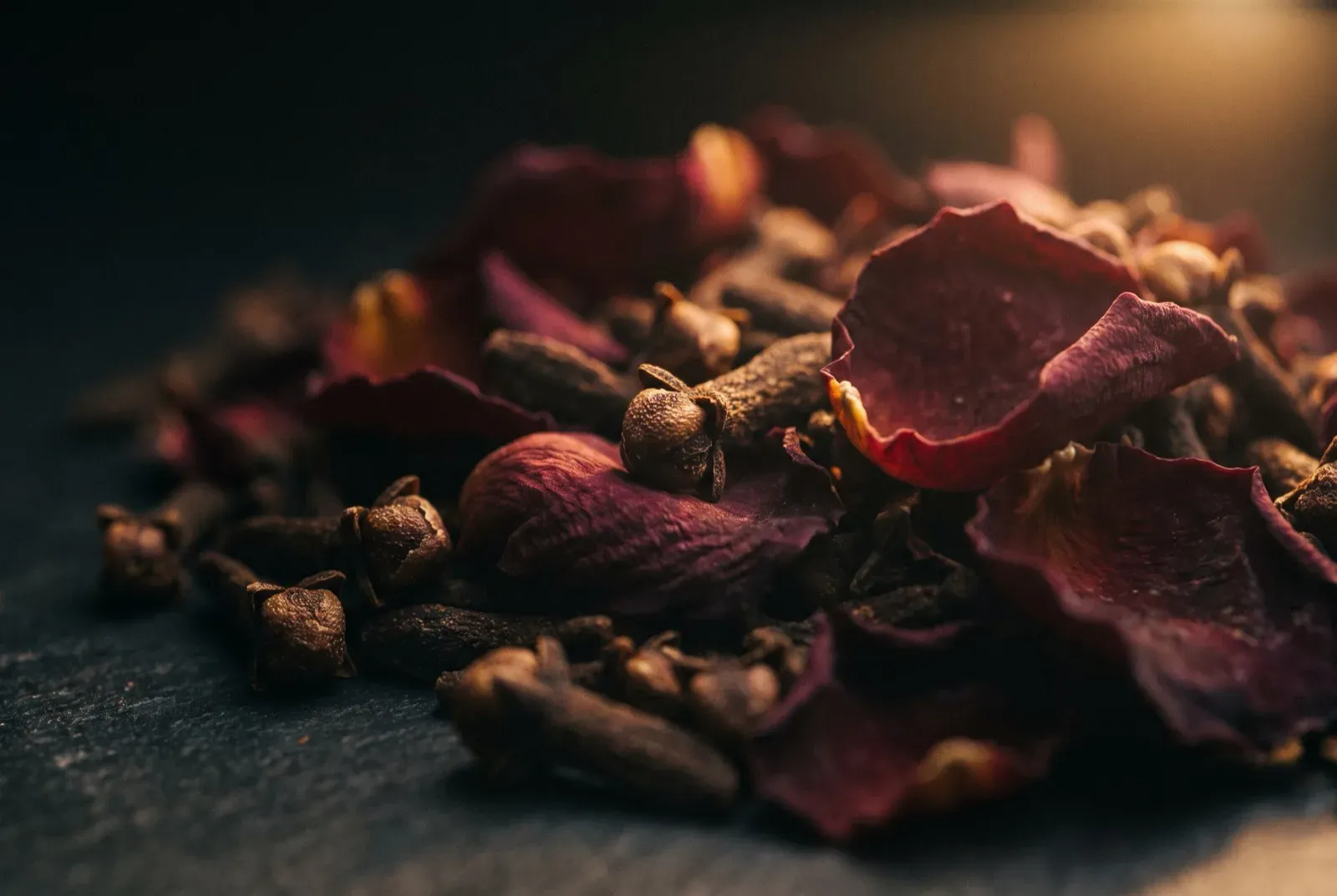 Whole cloves and dark dried rose petals layered in deep shadow, lit by a single warm light.